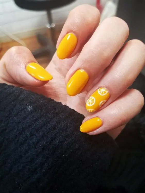 Rounded-shaped nails are painted in a bold, bright yellow. The ring finger is adorned with white lemon slice designs, adding a zesty and refreshing touch, perfect for summer festivities.