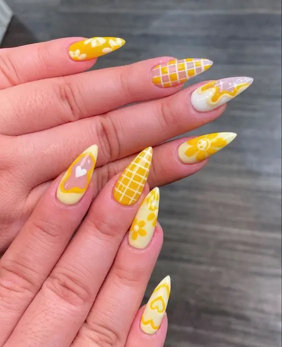 Stiletto nails in a mix of yellow, white, and pink, featuring various patterns like flowers, hearts, and grids. This fun and eclectic style captures the lively essence of summer.