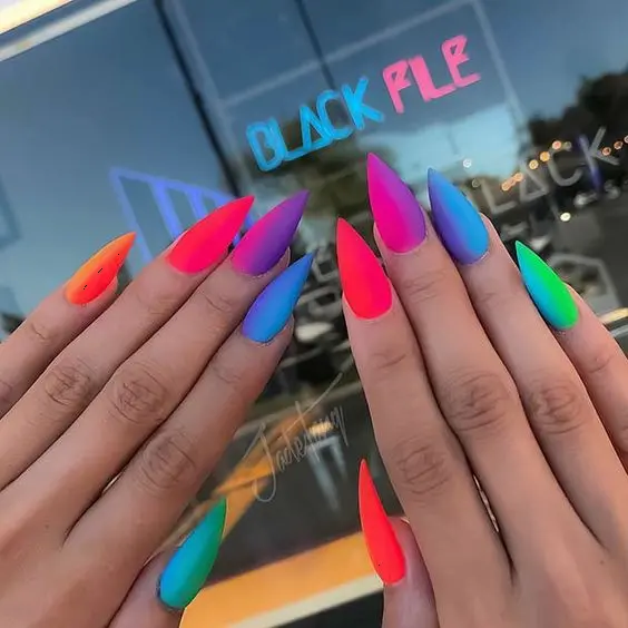 Matte stiletto nails display a striking neon rainbow gradient, blending from pink to purple to green. The matte finish adds a modern twist to the vibrant colors, making this design ideal for a trendy and fashion-forward style.