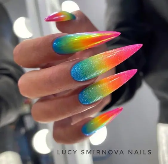 Long, stiletto-shaped nails are adorned with a glittery neon rainbow gradient, blending from blue to pink. The added shimmer enhances the vibrant colors, creating a glamorous and eye-catching look, ideal for making a bold fashion statement.