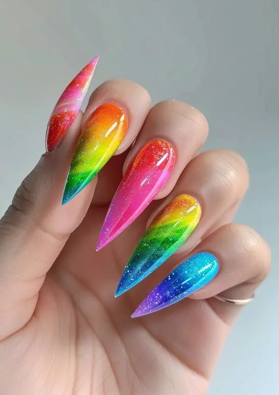 These nails feature a stunning neon rainbow gradient with glitter, transitioning from pink to blue on stiletto-shaped nails. The vibrant colors and sparkle create a dynamic and eye-catching look, perfect for a bold and playful personality.