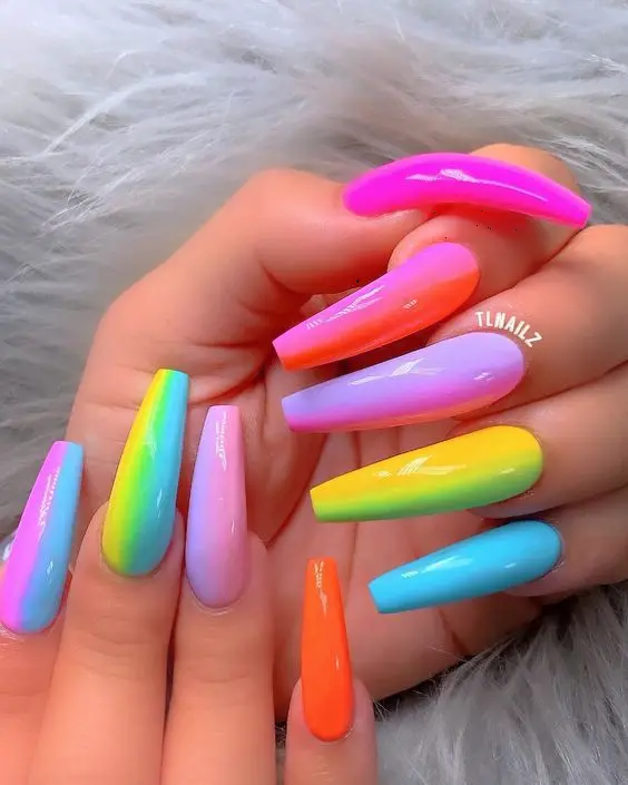 Long ballerina nails feature a vibrant neon gradient that flows from one nail to the next, creating a seamless rainbow effect. Each nail is painted with a different color, showcasing a glossy finish that adds to their charm and appeal.