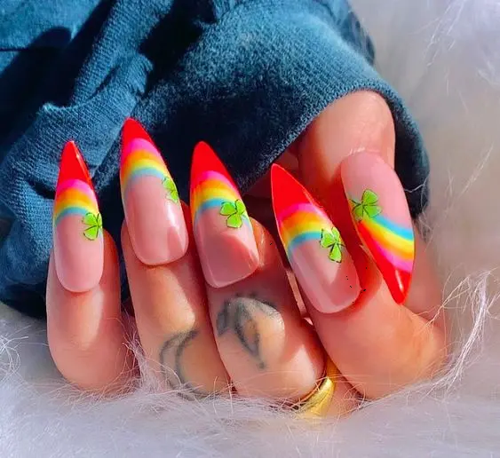 Long, stiletto nails are designed with a nude base, accented by a neon rainbow tip and small green shamrock decals. The sharp shape and playful design make these nails perfect for expressing a cheerful and lucky personality, ideal for festive occasions.