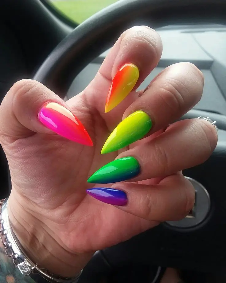 Stiletto-shaped nails exhibit a vivid neon rainbow gradient, transitioning from pink to green to blue. The bright colors and sleek form create a dynamic and bold style, perfect for someone who loves to stand out and make a statement.