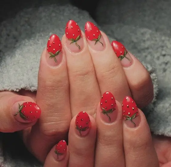 This unique design presents almond nails painted in solid red with a realistic strawberry pattern, complete with tiny green leaves and white specks mimicking seeds. The striking resemblance to actual strawberries makes this design fun and eye-catching.