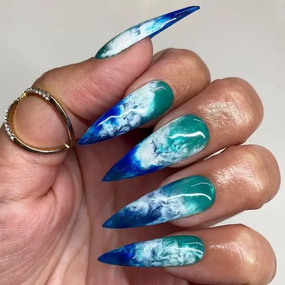 Dramatic stiletto nails featuring a deep blue to turquoise gradient with intricate wave designs. The bold colors and fluid patterns emulate the ocean, ideal for making a summer statement.