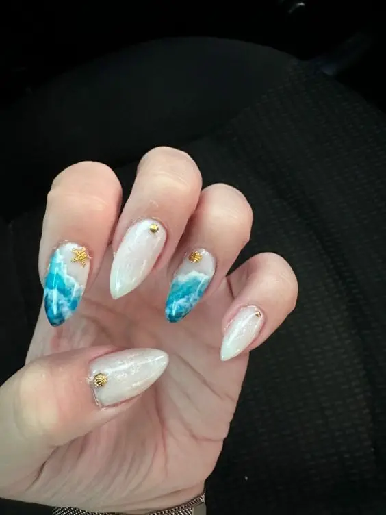 Stiletto-shaped nails adorned with blue and white wave patterns, complemented by gold starfish and seashell accents. This design captures the tranquility of the beach, making it perfect for summer adventures.