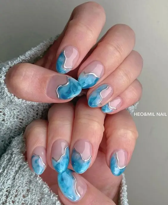 Featuring a short, almond shape, these nails depict a serene ocean wave design. The mix of light blue and white with marbling effects mimics the waves crashing on the shore. The calming color palette captures the tranquility of a summer day by the sea.