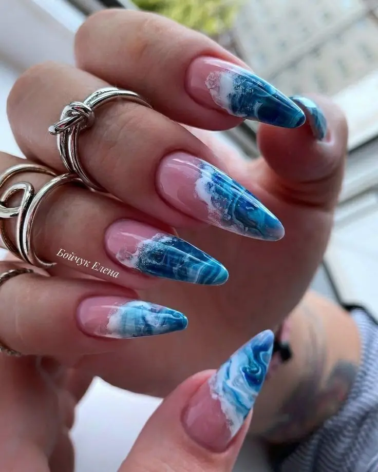 Stiletto nails with a translucent pink base and vivid blue ocean wave tips create a striking summer beach aesthetic. The detailed wave patterns bring a dynamic, summery vibe, ideal for seaside outings.