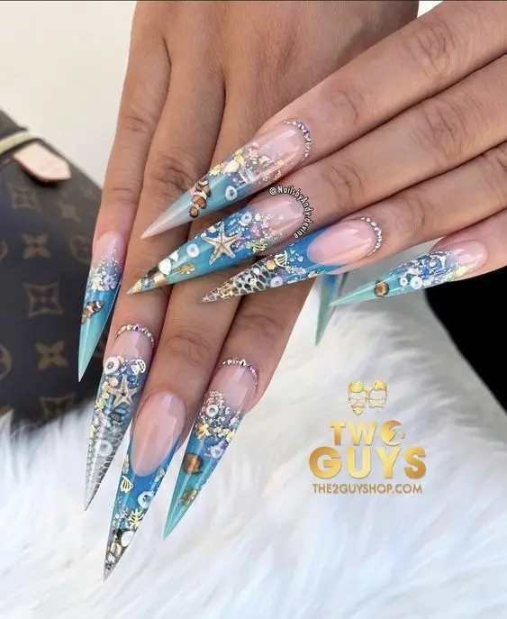 Long, pointed stiletto nails adorned with intricate sea-themed decorations, including starfish and seashells. The blue and nude gradient captures the essence of a summer beach with sparkling embellishments.