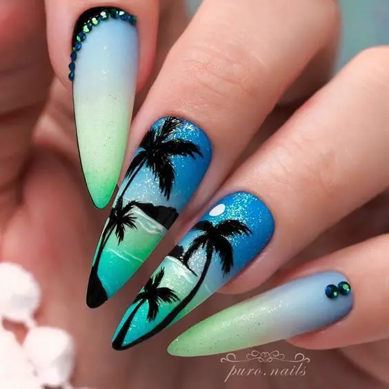 These stiletto nails highlight a vibrant beach sunset with intricate palm tree silhouettes. The gradient from blue to green with subtle sparkles adds a touch of tropical paradise, embodying summer nights.