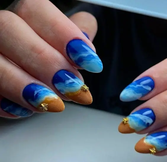 Almond-shaped nails with blue ocean waves and sandy tips. The design blends deep blues with sandy textures, creating a beachy, summer feel.