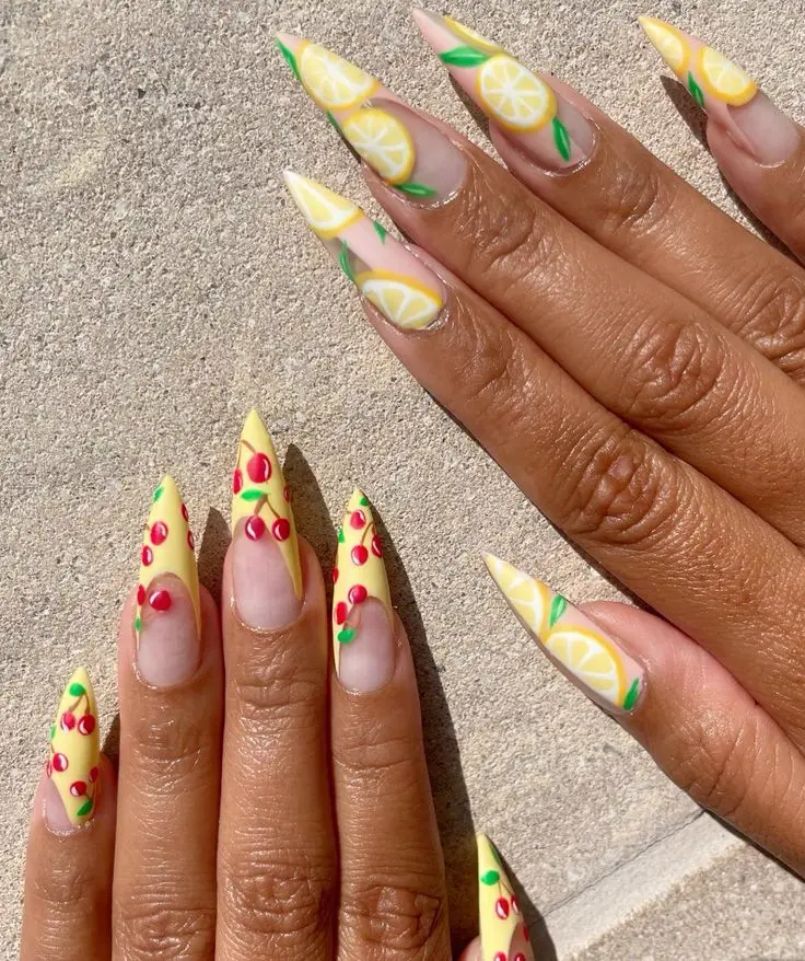 Long, pointed stiletto nails with a sunny yellow base are adorned with lemon and cherry designs, perfect for a playful and fruity summer look.