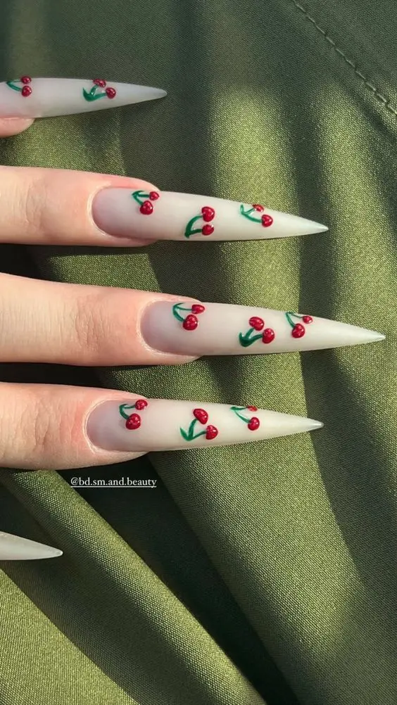 White stiletto nails adorned with cherry patterns, set against a soft, muted green background for a chic, summer-ready look.