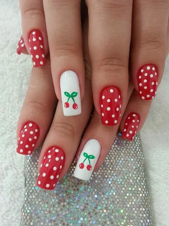 Bold red nails with white polka dots and cherry art on accent nails combine retro vibes with a touch of playful summer fruit.