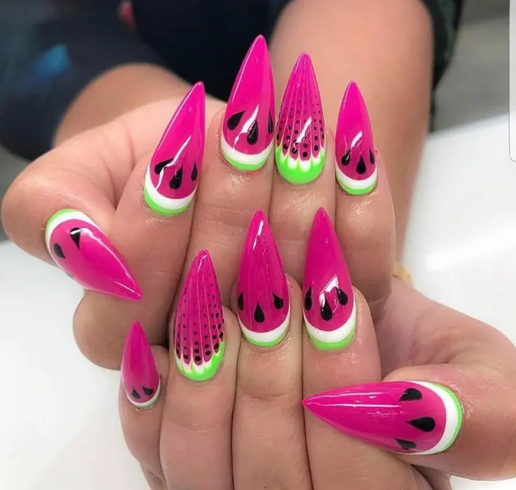 Stiletto nails with a juicy watermelon design are a bold summer statement, combining vibrant pink and green with black seed details.