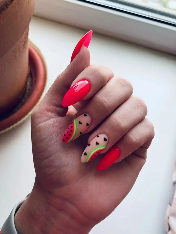 Sharp, stiletto-shaped nails in a bold red, with accent nails depicting watermelon slices, make for a strikingly vibrant summer statement.