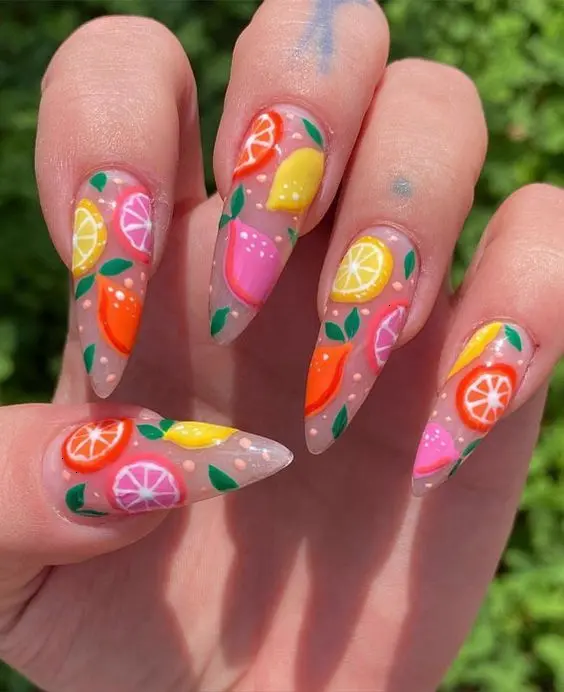 Translucent stiletto nails are a canvas for a colorful fruit salad design, mixing lemons, oranges, and strawberries for a bright summer feel.