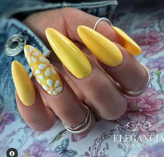 This summer nail design showcases long, almond-shaped nails painted in vibrant yellow. The accent nail features white and yellow daisies on a clear base, adding a touch of floral elegance to the bold, sunny look, ideal for summer.