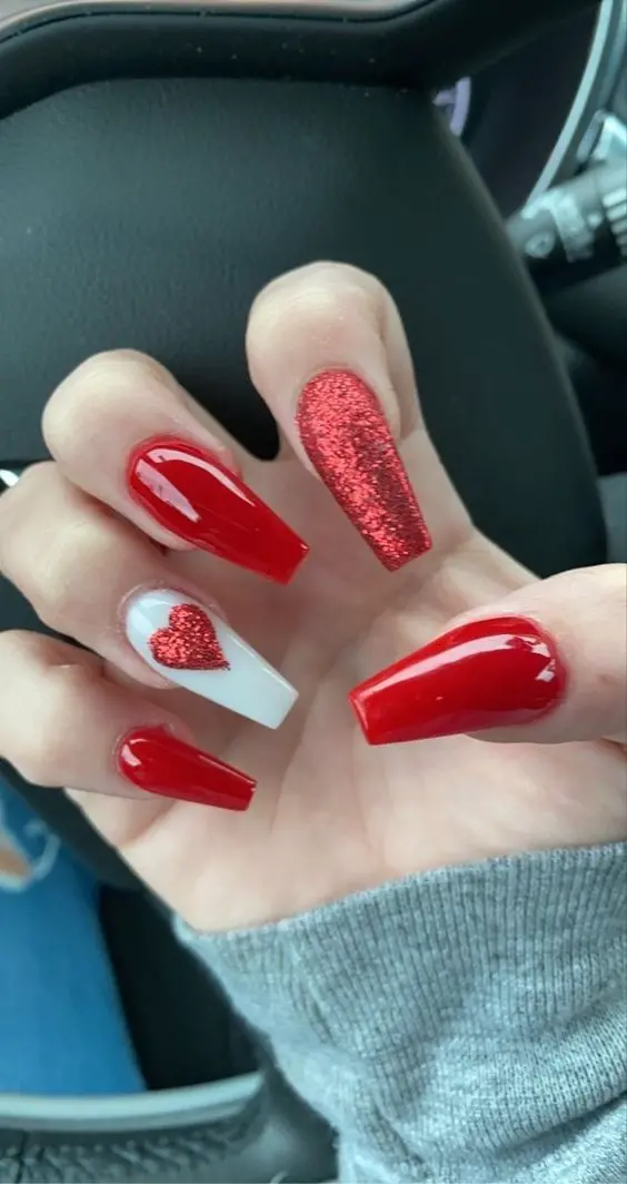 This bold design features glossy red polish with a glittery red accent nail and a white nail adorned with a red glitter heart. The combination of glossy and glitter finishes creates a glamorous and eye-catching look.