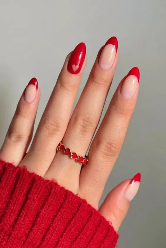 These elegant nails feature red French tips on a nude base, with one accent nail showcasing a small red heart. The almond shape and minimalist heart details offer a timeless and romantic look.