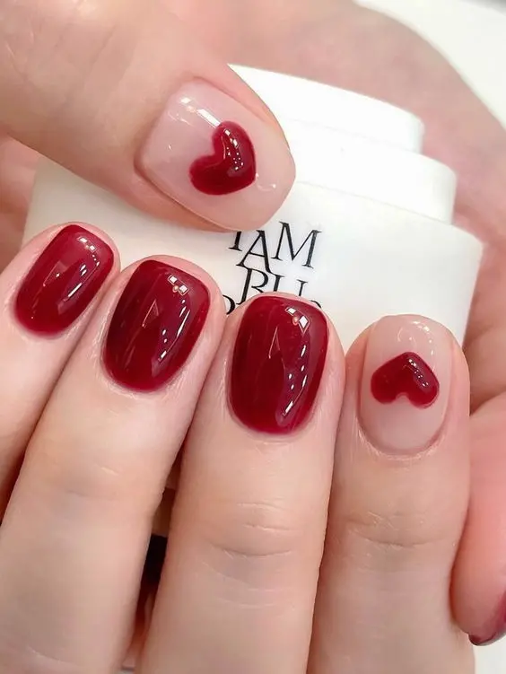 A trendy red heart nail design showcases a glossy maroon base with accent nails featuring red hearts. The squoval shape and high-shine finish add a touch of elegance, perfect for celebrating love and affection.