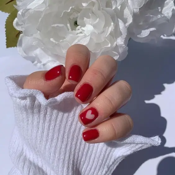 These bold square-shaped nails combine glossy red polish with a single white accent nail featuring a red heart. The striking color contrast and minimalist design create a stylish and modern look.