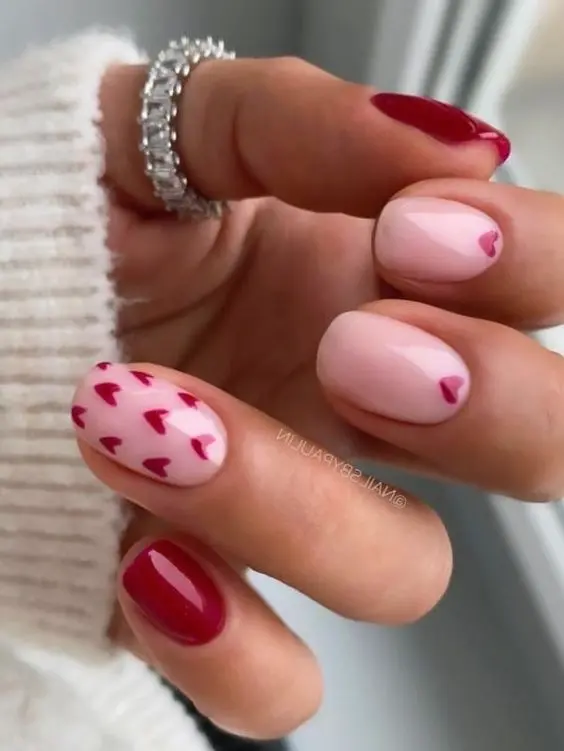 These short nails feature glossy red polish with accent nails adorned with small red hearts on a nude base. The playful design and vibrant colors make for a cute and charming look.