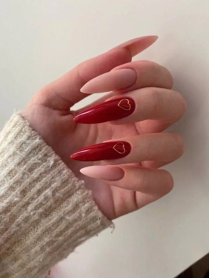 This design showcases long, stiletto-shaped nails with a mix of matte nude and glossy red polish. A single accent nail features a minimalist gold heart outline, adding a touch of elegance. This style is perfect for a sleek and modern look with a romantic twist.