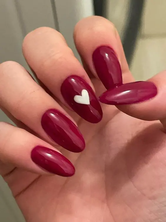 This sophisticated nail design features deep red polish on almond-shaped nails, with one nail adorned with a simple white heart. The rich red color and delicate heart accent convey elegance and a touch of romance.