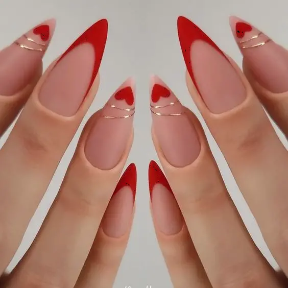 These stiletto nails boast a bold red French tip with delicate gold lines, accentuated by small red heart designs. The matte nude base enhances the vibrant red, creating a chic and sophisticated style ideal for making a statement.